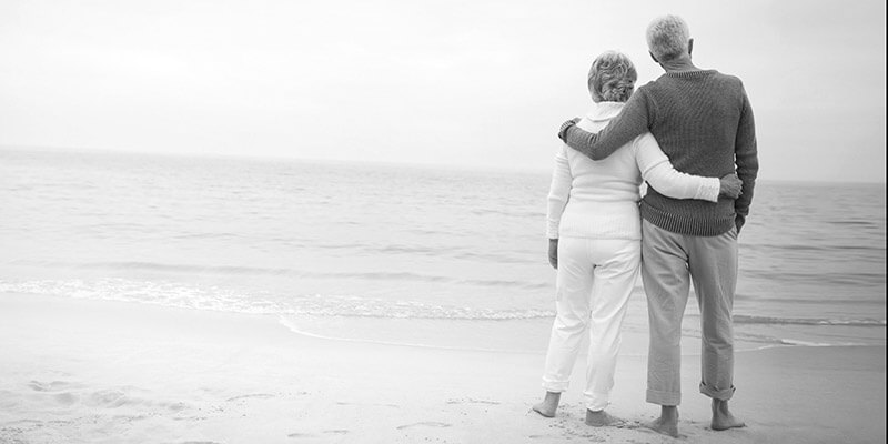 Retired couple barefoot in the sand stood looking out at the ocean